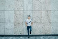 Young Asian man in casual wear leaning against a textured concrete wall with a confident expression.